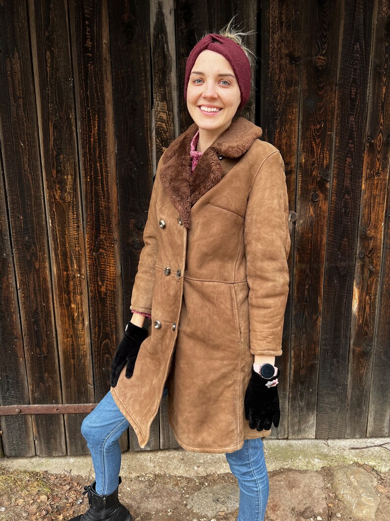 May include: A brown shearling coat with a double-breasted design. The coat features a sheepskin lining and a wide collar. The person in the image is wearing black gloves, blue jeans, and a burgundy headband. The background is a wooden door.