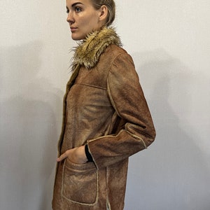 May include: A brown faux suede jacket featuring a tan faux fur collar, long sleeves, and two large front patch pockets. The jacket has visible tan faux fur trim along the edges and a straight hemline with side slits, worn over blue denim jeans.