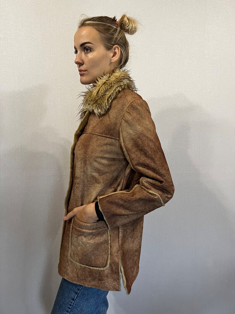 May include: A brown faux suede jacket featuring a tan faux fur collar, long sleeves, and two large front patch pockets. The jacket has visible tan faux fur trim along the edges and a straight hemline with side slits, worn over blue denim jeans.