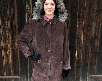 May include: A brown suede coat with a faux fur-lined hood. The coat has a button-down front, two front pockets, and gold-colored buttons. The person is wearing black gloves and blue jeans.