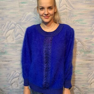 Può includere: Un maglione blu brillante con scollo tondo e maniche lunghe. Il maglione presenta un design intrecciato strutturato al centro e polsini a coste. Il materiale morbido e soffice crea un look accogliente ed elegante. Il maglione è indossato con jeans blu.