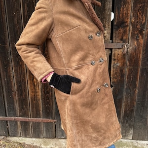 May include: A brown suede coat with a shearling collar and lining. The double-breasted coat features large, round buttons and a classic tailored design. The coat has two front pockets and is worn with blue jeans.