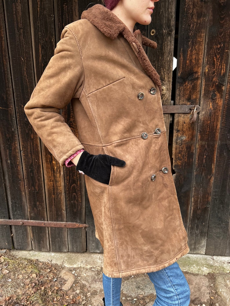 May include: A brown suede coat with a shearling collar and lining. The double-breasted coat features large, round buttons and a classic tailored design. The coat has two front pockets and is worn with blue jeans.