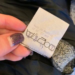 May include: A close-up of a clothing label with the text "Made in Austria" and washing instructions. The label is white with black text and symbols. The background is a black lining and a textured grey fabric.