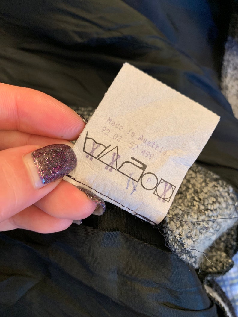 May include: A close-up of a clothing label with the text "Made in Austria" and washing instructions. The label is white with black text and symbols. The background is a black lining and a textured grey fabric.