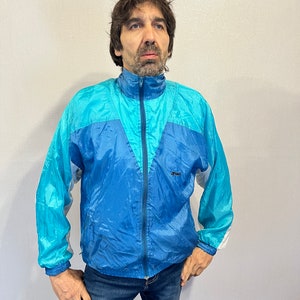 Puede incluir: Chaqueta de chándal vintage de nailon azul y turquesa con cremallera completa y cuello alzado. La chaqueta tiene detalles blancos en las mangas y un pequeño logotipo en el pecho. La persona lleva vaqueros azules.