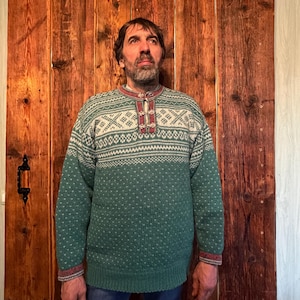 May include: A green and white patterned sweater with a round neck and a buttoned placket. The sweater features a geometric design on the yoke and cuffs. The sleeves and body are a solid green colour. The person is wearing blue jeans.