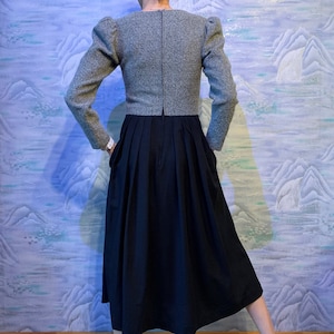 May include: A grey tweed jacket with puffed sleeves and a black pleated skirt. The jacket has a high neckline and long sleeves. The skirt is knee-length. The outfit is a vintage style.
