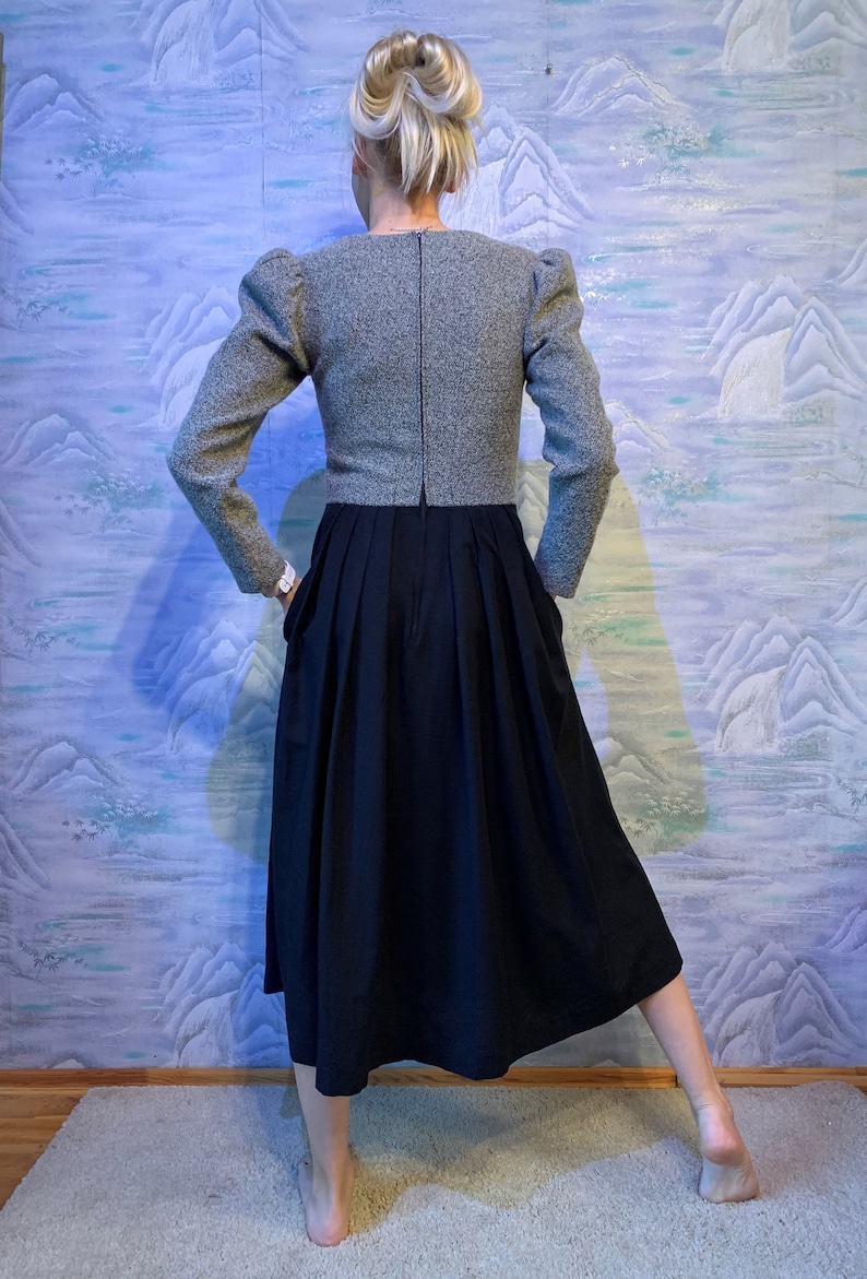 May include: A grey tweed jacket with puffed sleeves and a black pleated skirt. The jacket has a high neckline and long sleeves. The skirt is knee-length. The outfit is a vintage style.