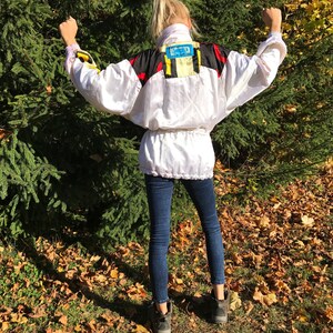 May include: A white vintage windbreaker jacket with black, red, and yellow accents. The jacket has a high collar and a cinched waist. The person is wearing dark blue jeans and black boots. The background is a green tree and fallen leaves.