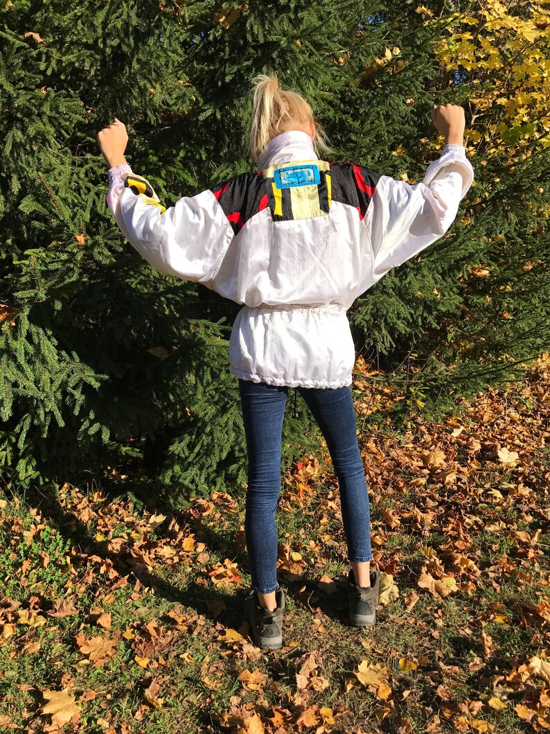 May include: A white vintage windbreaker jacket with black, red, and yellow accents. The jacket has a high collar and a cinched waist. The person is wearing dark blue jeans and black boots. The background is a green tree and fallen leaves.
