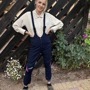 May include: A woman wearing a light beige button-down shirt with black braces and dark blue dungarees with purple accents. The dungarees have a zip down the front and knee pads.