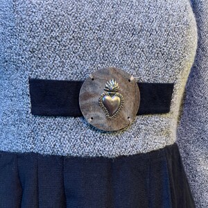 May include: A grey dress with a black waistband and skirt. The dress features a silver-toned metal embellishment with a heart design at the waist. The dress has long sleeves and a textured fabric.