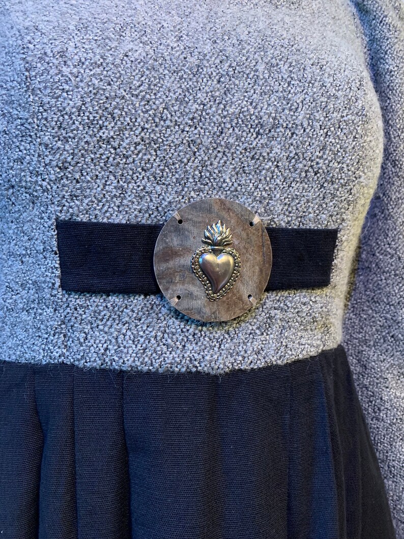 May include: A grey dress with a black waistband and skirt. The dress features a silver-toned metal embellishment with a heart design at the waist. The dress has long sleeves and a textured fabric.