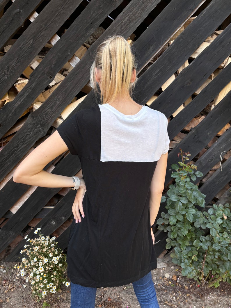 May include: A black and white t-shirt with a white panel on the back. The shirt has short sleeves and a relaxed fit.