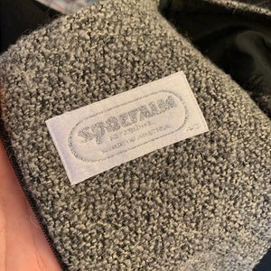 May include: Close-up of a grey and white textured fabric with a label that reads "SPARTAN" in silver lettering. The label also indicates "MADE IN AUSTRIA". The fabric appears to be part of a coat or jacket.