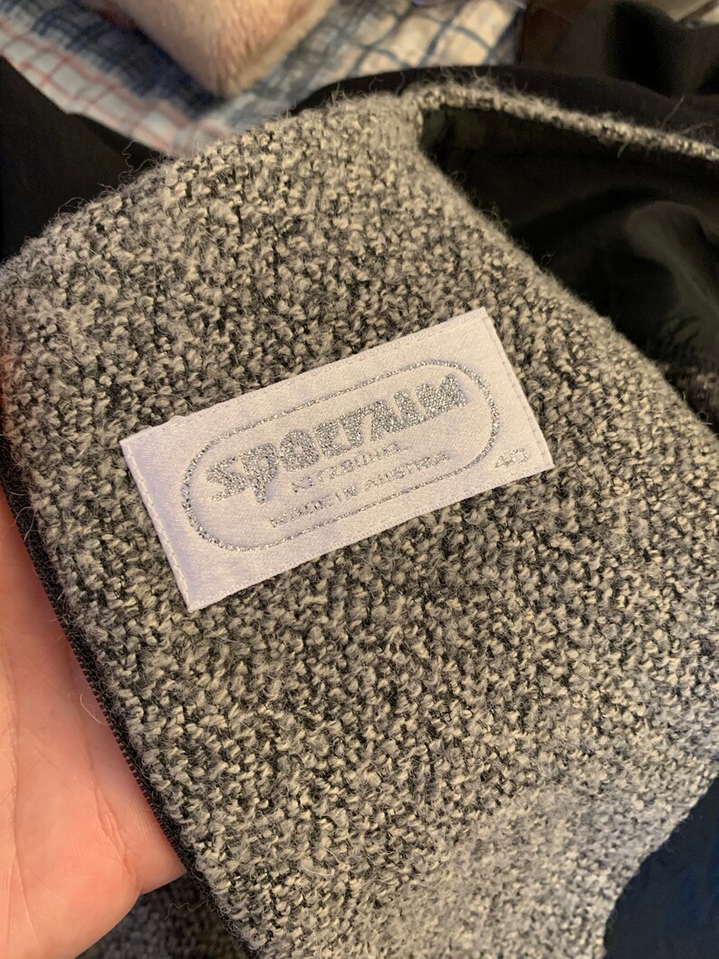 May include: Close-up of a grey and white textured fabric with a label that reads "SPARTAN" in silver lettering. The label also indicates "MADE IN AUSTRIA". The fabric appears to be part of a coat or jacket.