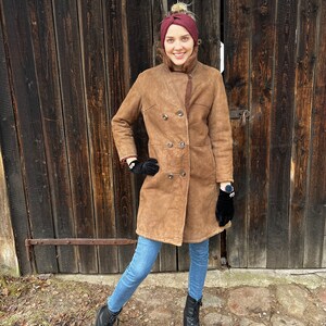 May include: A woman wearing a brown suede coat with a shearling lining, double-breasted buttons, and black gloves. She is also wearing a burgundy headband, blue jeans, and black lace-up boots. The background is a weathered wooden door.