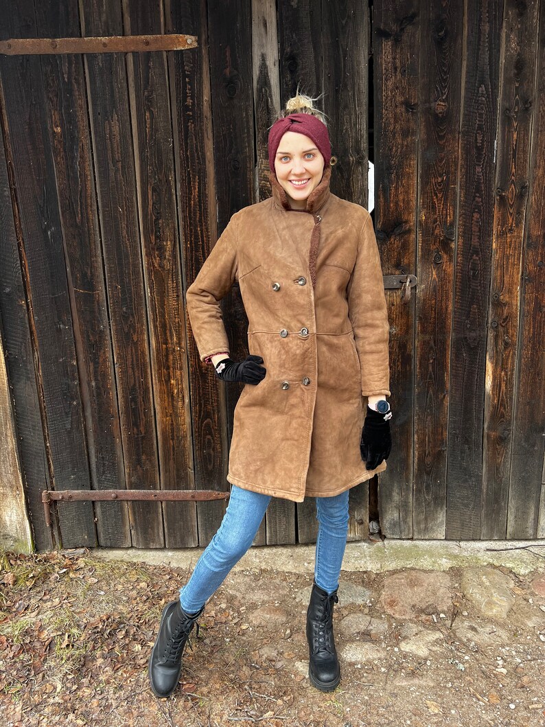 May include: A woman wearing a brown suede coat with a shearling lining, double-breasted buttons, and black gloves. She is also wearing a burgundy headband, blue jeans, and black lace-up boots. The background is a weathered wooden door.