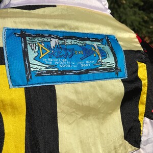 May include: A colorful jacket with a white collar and a blue label. The label has the text "DRESS THE BODY" and other text. The jacket has black, yellow, and white panels. The jacket is made of a shiny material.