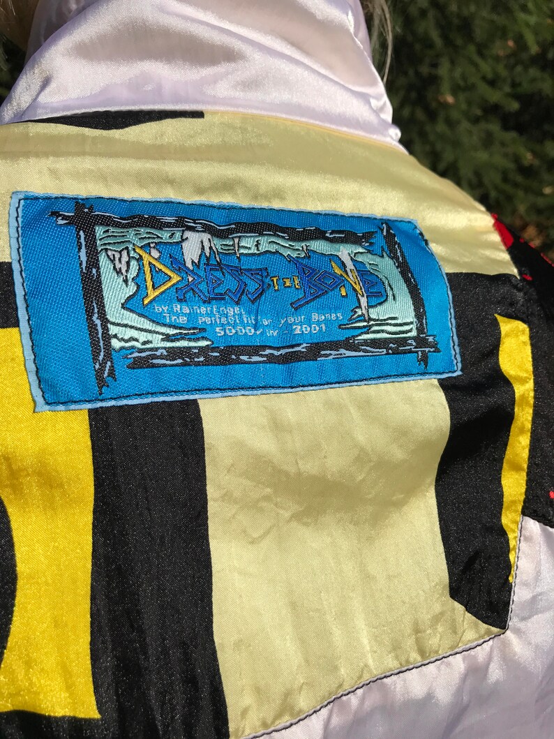 May include: A colorful jacket with a white collar and a blue label. The label has the text "DRESS THE BODY" and other text. The jacket has black, yellow, and white panels. The jacket is made of a shiny material.