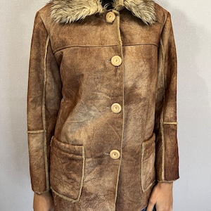 May include: A brown jacket with a distressed finish, featuring a large faux fur collar, four light-colored round buttons down the front, and two front patch pockets. The garment has visible seams along the sleeves and torso with a textured appearance.