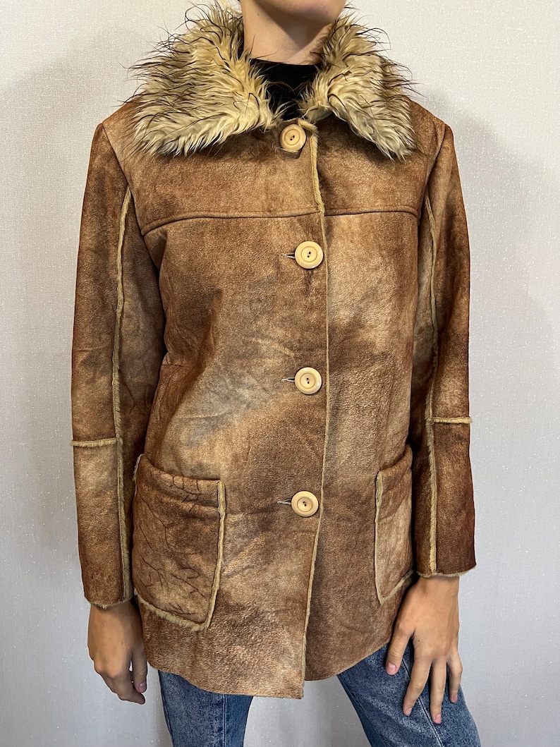 May include: A brown jacket with a distressed finish, featuring a large faux fur collar, four light-colored round buttons down the front, and two front patch pockets. The garment has visible seams along the sleeves and torso with a textured appearance.