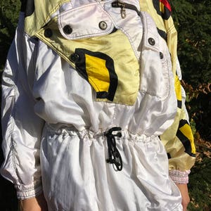 May include: A vintage white and yellow windbreaker jacket with black and red accents. The jacket features a high collar, a zipper closure, and a drawstring waist. The design includes black and yellow geometric shapes.