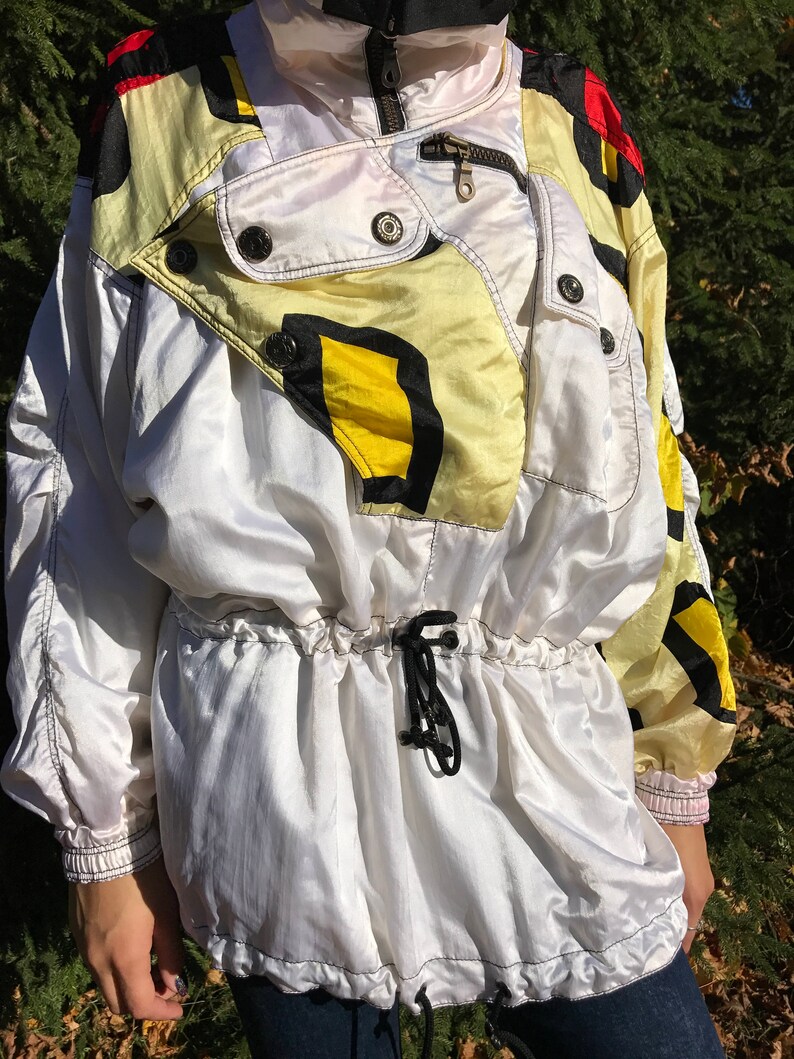 May include: A vintage white and yellow windbreaker jacket with black and red accents. The jacket features a high collar, a zipper closure, and a drawstring waist. The design includes black and yellow geometric shapes.