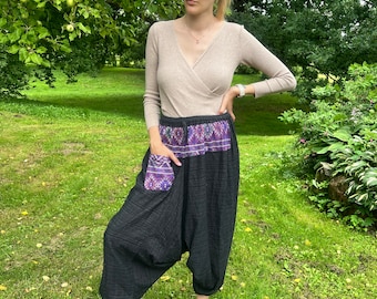 May include: Black harem pants with a purple and white patterned waistband and pocket. The pants have a loose, comfortable fit. The model is wearing a beige long-sleeve top and black sneakers.
