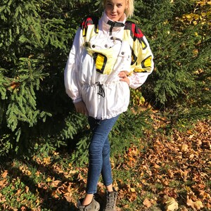 May include: White and yellow vintage ski jacket with black and red accents. The jacket has a high collar, a drawstring waist, and multiple pockets. The model is wearing dark blue jeans and gray hiking boots.