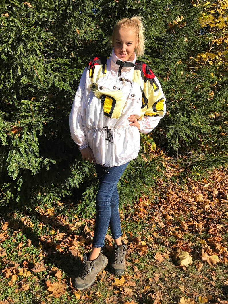 May include: White and yellow vintage ski jacket with black and red accents. The jacket has a high collar, a drawstring waist, and multiple pockets. The model is wearing dark blue jeans and gray hiking boots.