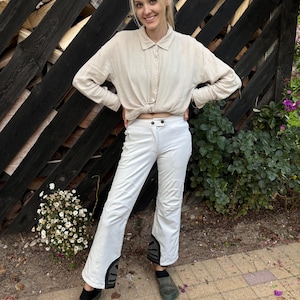 May include: A woman wearing a light beige button-down shirt tied at the waist, white pants with black accents, and dark green shoes. She is standing in front of a wooden fence.