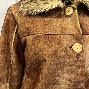 May include: A brown suede jacket featuring a cream-colored faux fur collar and trim along the seams. The jacket is fastened with three large, round, light beige buttons down the front, showcasing a textured finish and visible stitching details.