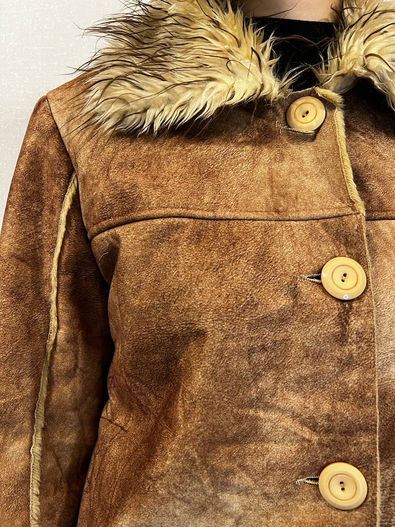 May include: A brown suede jacket featuring a cream-colored faux fur collar and trim along the seams. The jacket is fastened with three large, round, light beige buttons down the front, showcasing a textured finish and visible stitching details.