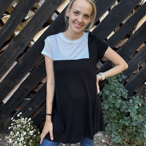 May include: A woman wearing a black and white t-shirt dress with short sleeves and blue jeans. She is standing in front of a wooden fence.