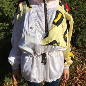 May include: A vintage white and yellow windbreaker jacket with a high collar and black zipper. The jacket features a drawstring waist and a colorful geometric pattern on the shoulders. The sleeves have elastic cuffs.