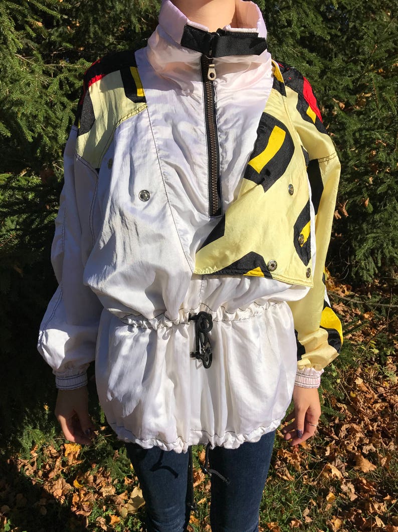 May include: A vintage white and yellow windbreaker jacket with a high collar and black zipper. The jacket features a drawstring waist and a colorful geometric pattern on the shoulders. The sleeves have elastic cuffs.