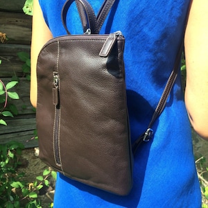 May include: A brown leather backpack with a vertical zipper and white stitching. The backpack has adjustable straps and a top handle. The person in the image is wearing a blue dress.