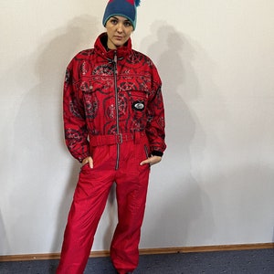 Puede incluir: Traje de esquí rojo y negro vintage con una parte superior estampada y pantalones rojos lisos. El traje tiene cremallera, cinturón y un pequeño parche con el logo. Un gorro con pompón azul, rojo y turquesa completa el conjunto. El tejido es brillante.