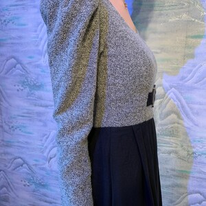 May include: A grey and black dress with a V-neck and puffed sleeves. The top of the dress is grey with a textured pattern, and the sleeves are long and puffed. The bottom of the dress is black and pleated, with a belt at the waist.