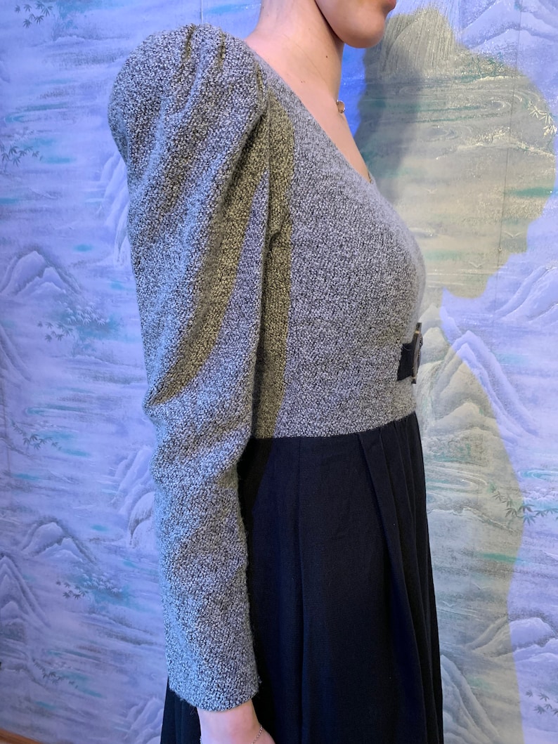 May include: A grey and black dress with a V-neck and puffed sleeves. The top of the dress is grey with a textured pattern, and the sleeves are long and puffed. The bottom of the dress is black and pleated, with a belt at the waist.