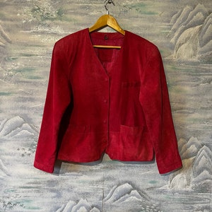 Peut inclure: Veste en daim rouge avec un col en V, une fermeture boutonnée et deux poches avant. La veste est suspendue à un cintre en bois sur un fond de papier peint.