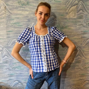 May include: A blue and white gingham top with a square neckline and short, puffed sleeves. The top has a row of buttons down the front and is paired with blue jeans. The top has white lace trim.