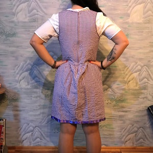 May include: A vintage-style dress featuring a white top and a checkered purple and white bodice. The dress has a fitted waist and a flared skirt with a purple ribbon trim. The dress is worn over a white short-sleeved shirt.