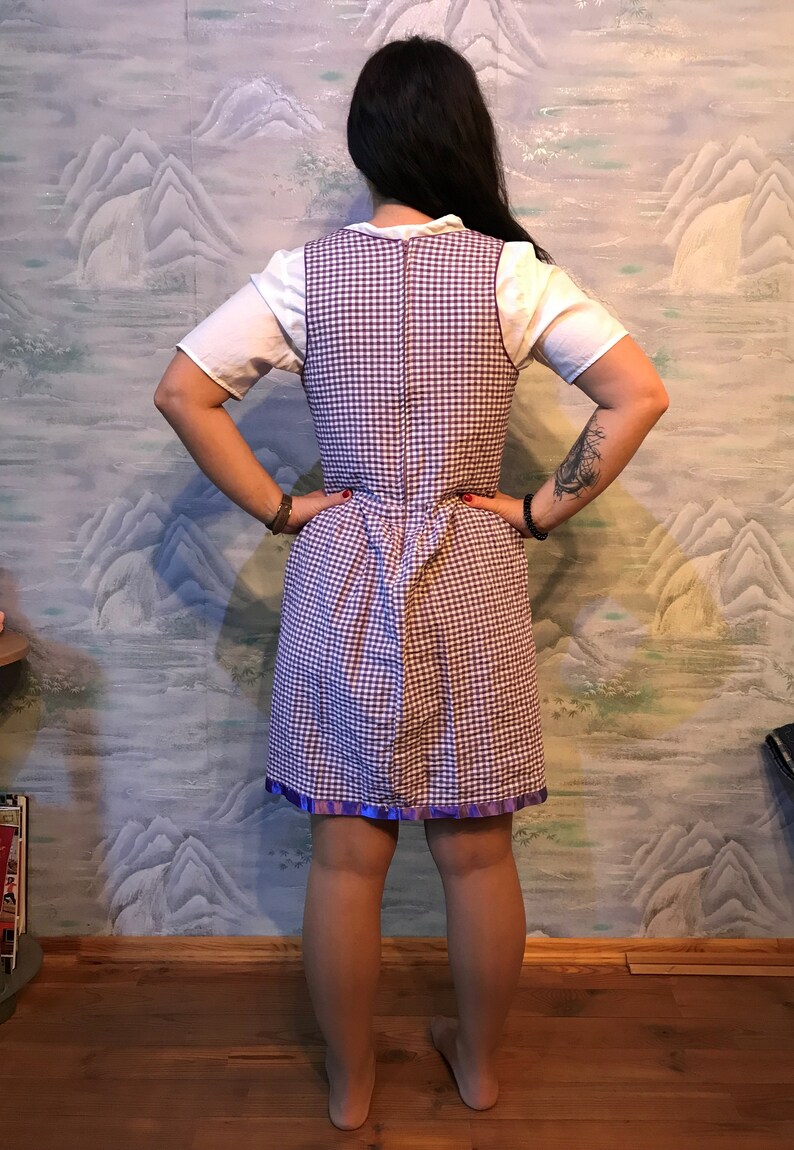 May include: A vintage-style dress featuring a white top and a checkered purple and white bodice. The dress has a fitted waist and a flared skirt with a purple ribbon trim. The dress is worn over a white short-sleeved shirt.