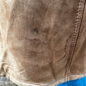 May include: Close-up of a brown suede jacket. The jacket has a textured surface and visible stitching along the seams. The bottom hem is curved. There is a small dark mark on the jacket's surface.