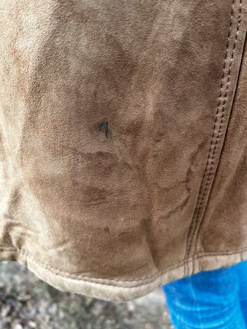 May include: Close-up of a brown suede jacket. The jacket has a textured surface and visible stitching along the seams. The bottom hem is curved. There is a small dark mark on the jacket's surface.