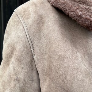 May include: Close-up of a tan suede jacket with a brown shearling collar. The jacket features visible stitching along the shoulder seam. The texture of the suede is soft and slightly worn, suggesting a vintage or well-loved piece.