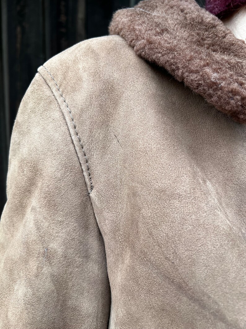May include: Close-up of a tan suede jacket with a brown shearling collar. The jacket features visible stitching along the shoulder seam. The texture of the suede is soft and slightly worn, suggesting a vintage or well-loved piece.